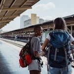 Two diverse female backpackers wait on a sunny railway platform, ready for adventure.